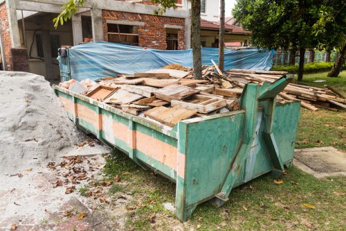 A large green metal skip filled with wooden debris, including planks and broken panels, situated on a grassy area next to a residential street. The skip is positioned on a patch of earth with some scattered leaves and debris on the ground nearby. In the background, there is a house under construction, partially covered with blue tarpaulin, and a tree with green foliage providing partial shade. The scene suggests a rubbish removal service in North Watford, with the skip used for clearing construction or renovation waste as part of a house clearance project managed by House Clearance North Watford. The environment appears calm and residential, with natural daylight illuminating the scene and the focus on the skip's contents and surrounding outdoor setting.