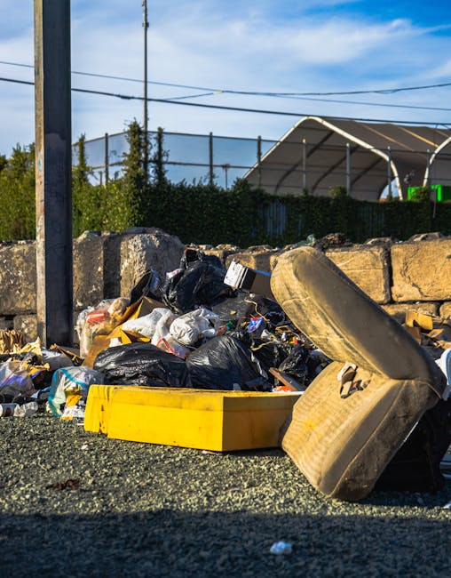 A small, old blue pickup truck parked on a street with its cargo bed fully loaded with assorted waste materials, including flattened cardboard boxes, black plastic garbage bags, and various other disc