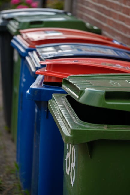 Close-up image of multiple black plastic rubbish bags, tied securely at the top, stacked against a plain, light-colored wall. The bags are made of shiny, slightly crinkled plastic material, with some